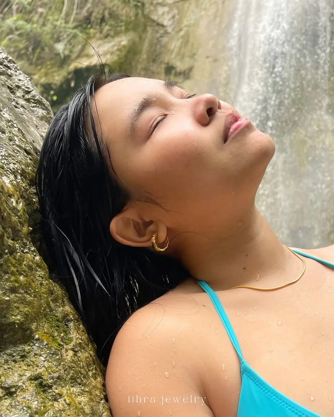 A woman by a waterfall, wet hair, wearing gold hoops and a necklace